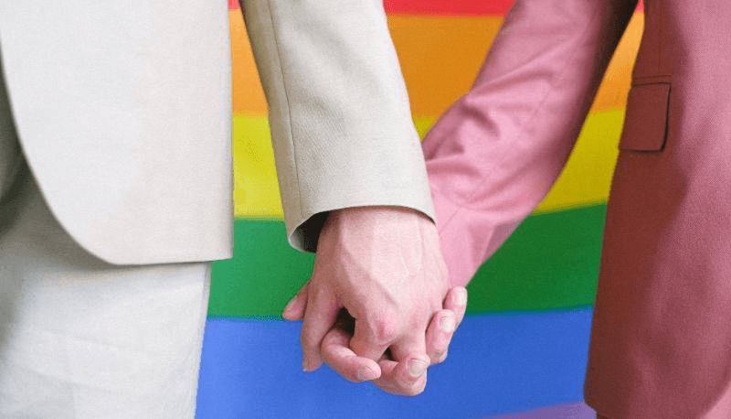 Discriminati per l'orientamento sessuale, due donne in tailleur si tengono la mano davanti a una bandiera arcobaleno