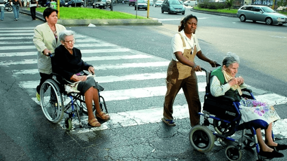 Badanti donne attraversano la strada con anziane in carrozzina