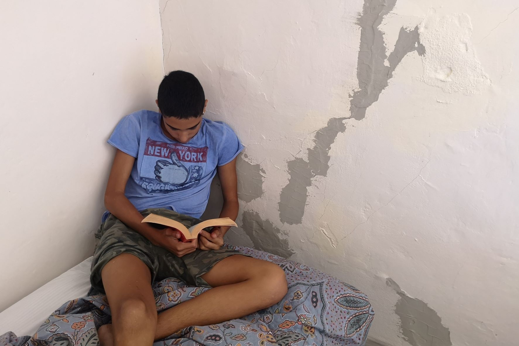 Giovani e povertà: un ragazzo seduto per terra legge un libro contro un muro scrostato.