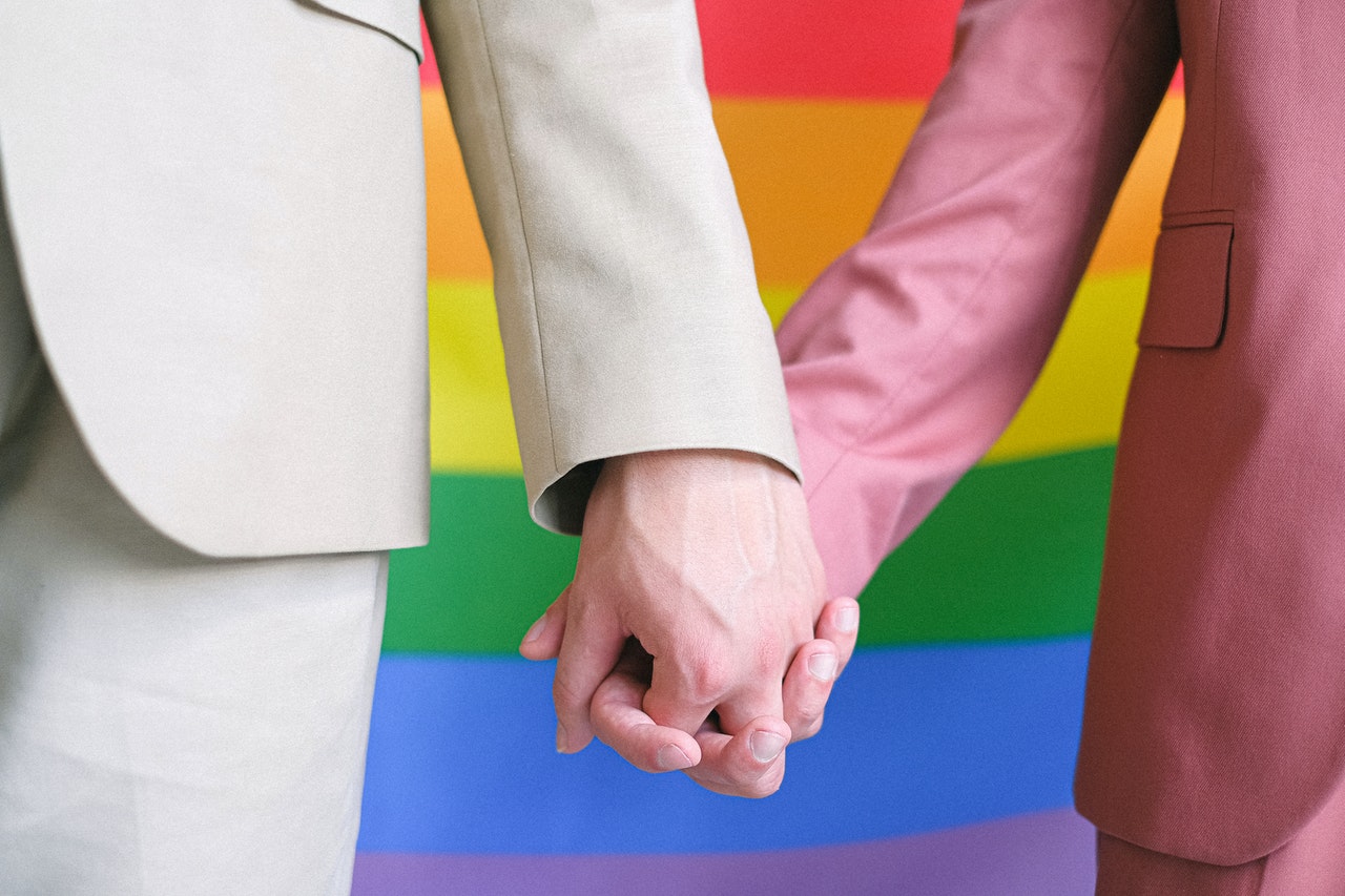 Orientamento sessuale nella PA: due donne in tailleur si tengono la mano con una bandiera arcobaleno sullo sfondo
